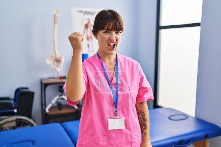 Téléchargez les photos : Young brunette woman working at rehabilitation clinic angry and mad raising fist frustrated and furious while shouting with anger. rage and aggressive concept. - en image libre de droit