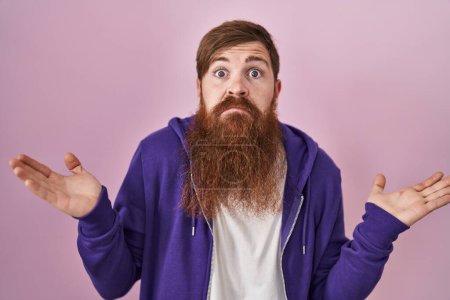 Photo for Caucasian man with long beard standing over pink background clueless and confused expression with arms and hands raised. doubt concept. - Royalty Free Image
