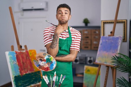 Photo for Brazilian young man holding painter palette at artist studio puffing cheeks with funny face. mouth inflated with air, catching air. - Royalty Free Image