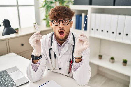 Photo for Arab doctor man with beard holding covid test in shock face, looking skeptical and sarcastic, surprised with open mouth - Royalty Free Image