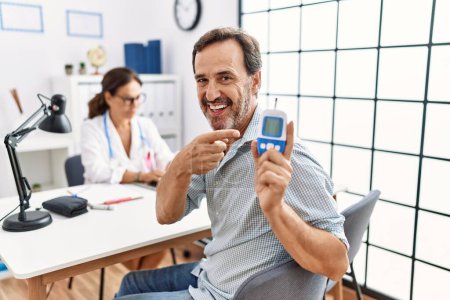 Téléchargez les photos : Middle age man at doctor clinic holding glucose meter device smiling happy pointing with hand and finger - en image libre de droit