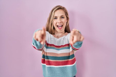 Photo for Young blonde woman standing over pink background pointing to you and the camera with fingers, smiling positive and cheerful - Royalty Free Image