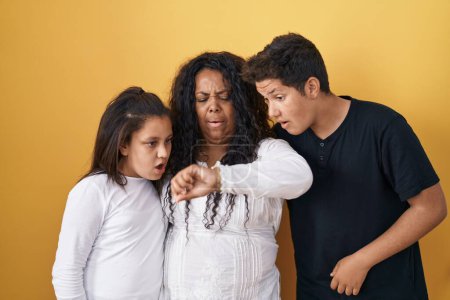 Téléchargez les photos : Famille de mère, fille et fils debout sur fond jaune regardant le temps de la montre inquiet, peur de se faire tard - en image libre de droit