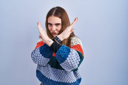 Foto de Young hispanic girl standing over blue background rejection expression crossing arms doing negative sign, angry face - Imagen libre de derechos
