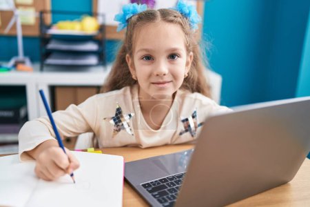 Foto de Young little girl sitting on the table doing homework with laptop looking positive and happy standing and smiling with a confident smile showing teeth - Imagen libre de derechos
