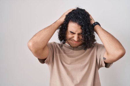 Foto de Hispanic man with curly hair standing over white background suffering from headache desperate and stressed because pain and migraine. hands on head. - Imagen libre de derechos