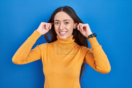 Téléchargez les photos : Jeune femme brune debout sur fond bleu souriant tirant les oreilles avec les doigts, geste drôle. problème d'audition - en image libre de droit