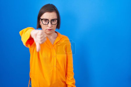 Foto de Middle age hispanic woman wearing glasses standing over blue background looking unhappy and angry showing rejection and negative with thumbs down gesture. bad expression. - Imagen libre de derechos