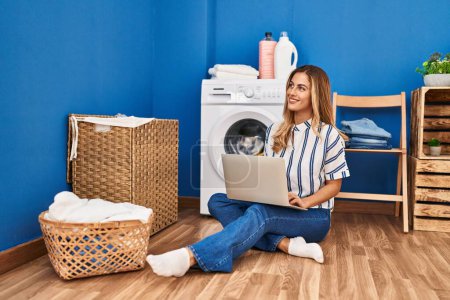 Photo for Young blonde woman using laptop waiting for washing machine at laundry room - Royalty Free Image