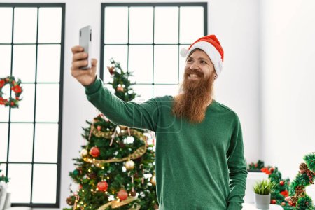 Téléchargez les photos : Jeune rousse homme faire selfie par le smartphone debout près de l'arbre de Noël à la maison - en image libre de droit