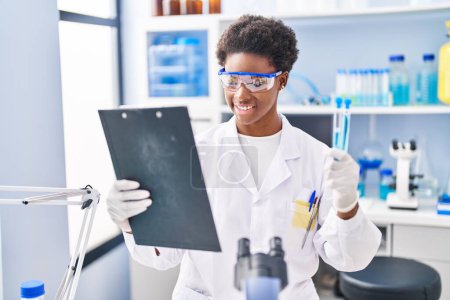 Téléchargez les photos : African american woman wearing scientist uniform reading document holding test tubes at laboratory - en image libre de droit