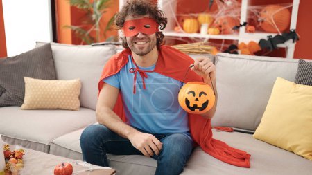Photo for Young hispanic man wearing superhero costume holding halloween pumpkin basket at home - Royalty Free Image