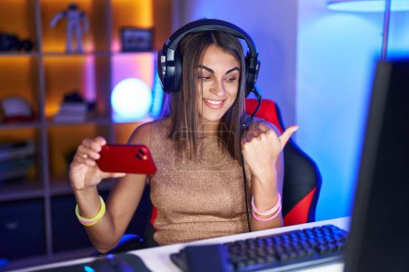 Téléchargez les photos : Young hispanic woman playing video games with smartphone pointing thumb up to the side smiling happy with open mouth - en image libre de droit