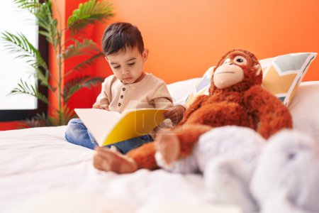 Foto de Adorable niño hispano leyendo libro sentado en la cama en el dormitorio - Imagen libre de derechos