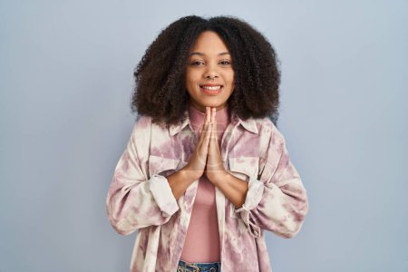 Foto de Young african american woman standing over blue background praying with hands together asking for forgiveness smiling confident. - Imagen libre de derechos