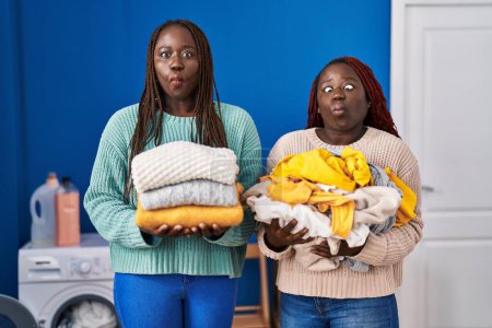 Foto de Dos mujeres africanas sosteniendo la ropa doblada después de planchar haciendo cara de pez con boca y ojos entrecerrados, loco y cómico. - Imagen libre de derechos