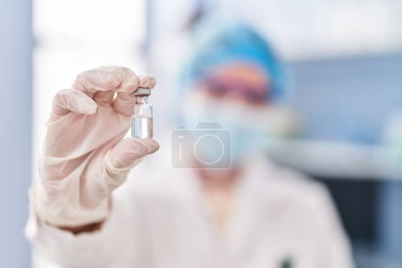 Photo for Young blonde woman wearing scientist uniform and medical mask holding covid-19 vaccine dose at laboratory - Royalty Free Image