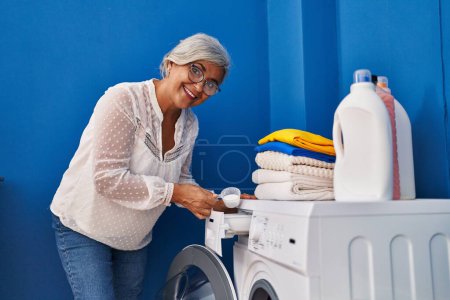 Foto de Mujer de mediana edad vertiendo detergente en la lavadora en la sala de lavandería - Imagen libre de derechos