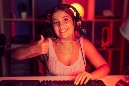 Foto de Young blonde woman playing video games wearing headphones doing happy thumbs up gesture with hand. approving expression looking at the camera showing success. - Imagen libre de derechos