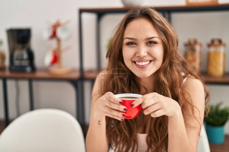 Photo for Young beautiful hispanic woman drinking coffee sitting on table at home - Royalty Free Image