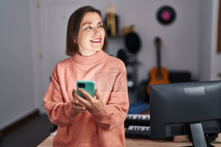 Foto de Middle age woman musician smiling confident using smartphone at music studio - Imagen libre de derechos