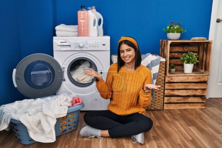 Téléchargez les photos : Young hispanic woman doing laundry smiling cheerful offering hands giving assistance and acceptance. - en image libre de droit