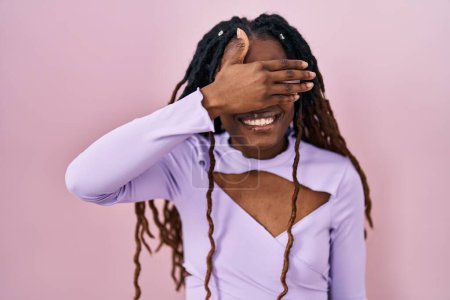 Foto de Mujer africana con el pelo trenzado de pie sobre fondo rosa sonriendo y riendo con la mano en la cara cubriendo los ojos para sorpresa. concepto ciego. - Imagen libre de derechos