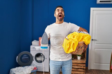 Foto de Joven hombre hispano con barba sosteniendo hierro y ropa en casa enojado y loco gritando frustrado y furioso, gritando con ira mirando hacia arriba. - Imagen libre de derechos