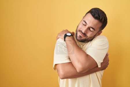 Photo for Handsome hispanic man standing over yellow background hugging oneself happy and positive, smiling confident. self love and self care - Royalty Free Image