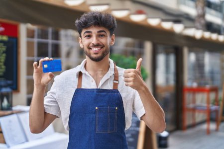 Téléchargez les photos : Arabe homme avec barbe portant tablier de serveur à la terrasse du restaurant tenant carte de crédit souriant heureux et positif, pouce levé faire excellent et signe d'approbation - en image libre de droit