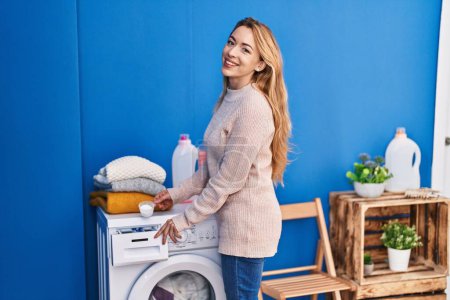 Téléchargez les photos : Jeune femme souriante confiante détergent versant sur la machine à laver à la buanderie - en image libre de droit