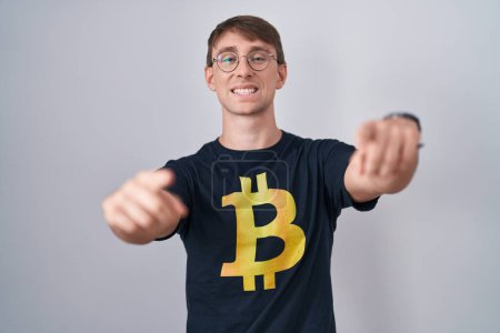 Téléchargez les photos : Homme blond caucasien portant Bitcoin t-shirt pointant vers vous et la caméra avec les doigts, souriant positif et joyeux - en image libre de droit