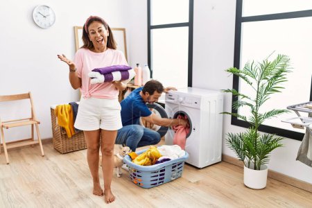 Photo for Middle age couple holding clean laundry celebrating achievement with happy smile and winner expression with raised hand - Royalty Free Image