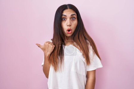 Photo for Young arab woman standing over pink background surprised pointing with hand finger to the side, open mouth amazed expression. - Royalty Free Image