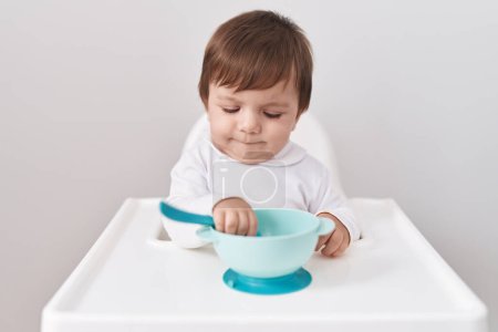 Photo for Adorable hispanic toddler sitting on highchair holding bowl over isolated white background - Royalty Free Image