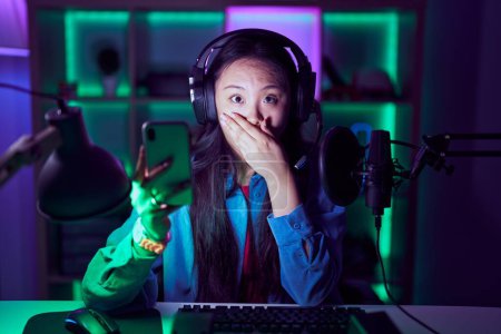 Téléchargez les photos : Young asian woman playing video games with smartphone shocked covering mouth with hands for mistake. secret concept. - en image libre de droit