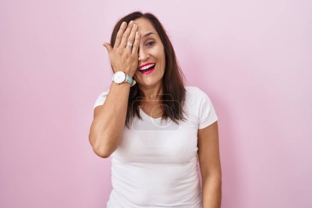 Photo for Middle age brunette woman standing over pink background covering one eye with hand, confident smile on face and surprise emotion. - Royalty Free Image