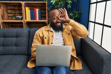 Foto de African american man using laptop at home sitting on the sofa smiling happy doing ok sign with hand on eye looking through fingers - Imagen libre de derechos