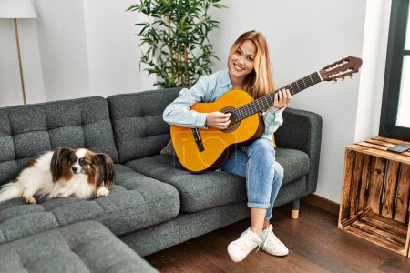 Foto de Joven mujer caucásica tocando guitarra clásica sentada en sofá con perro en casa - Imagen libre de derechos