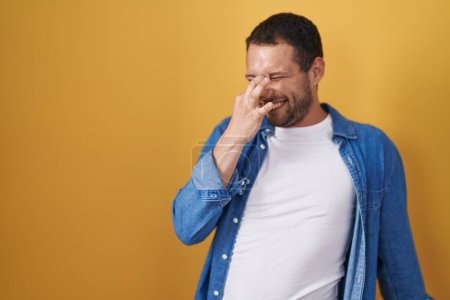 Foto de Hispanic man standing over yellow background smelling something stinky and disgusting, intolerable smell, holding breath with fingers on nose. bad smell - Imagen libre de derechos