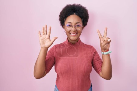 Foto de Hermosa mujer africana con el pelo rizado de pie sobre fondo rosa mostrando y apuntando hacia arriba con los dedos número siete mientras sonríe confiado y feliz. - Imagen libre de derechos
