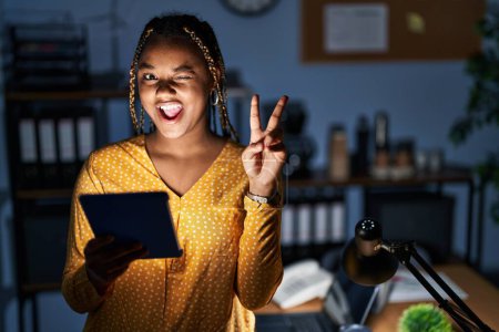 Téléchargez les photos : Femme afro-américaine avec des tresses travaillant au bureau la nuit avec une tablette souriant avec un sourire heureux clin d'oeil à la caméra faisant signe de victoire avec les doigts. numéro deux. - en image libre de droit