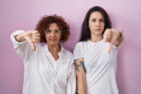 Photo for Hispanic mother and daughter wearing casual white t shirt over pink background looking unhappy and angry showing rejection and negative with thumbs down gesture. bad expression. - Royalty Free Image