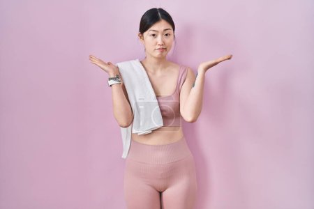 Foto de Chinese young woman wearing sportswear and towel clueless and confused expression with arms and hands raised. doubt concept. - Imagen libre de derechos