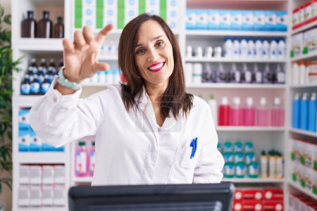 Téléchargez les photos : Middle age brunette woman working at pharmacy drugstore smiling and confident gesturing with hand doing small size sign with fingers looking and the camera. measure concept. - en image libre de droit