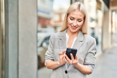 Téléchargez les photos : Jeune femme blonde souriant confiant en utilisant smartphone à la rue - en image libre de droit