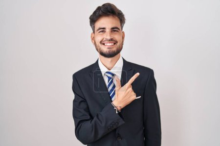 Foto de Young hispanic man with tattoos wearing business suit and tie cheerful with a smile on face pointing with hand and finger up to the side with happy and natural expression - Imagen libre de derechos