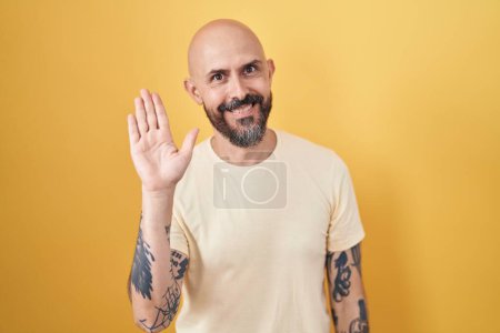 Téléchargez les photos : Hispanic man with tattoos standing over yellow background waiving saying hello happy and smiling, friendly welcome gesture - en image libre de droit