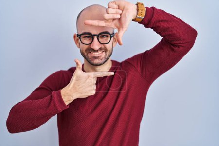 Foto de Young bald man with beard standing over white background wearing glasses smiling making frame with hands and fingers with happy face. creativity and photography concept. - Imagen libre de derechos