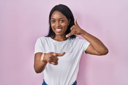 Téléchargez les photos : Jeune femme africaine portant t-shirt blanc décontracté souriant faire parler au téléphone geste et vous pointant du doigt. Appelle-moi. - en image libre de droit